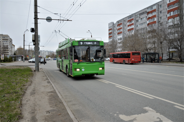 Ein grüner Bus fährt auf einer Stadtstraße mit hohen Gebäuden, Gras und Strommasten auf der linken Seite, Bäumen im Hintergrund und einem klaren blauen Himmel.