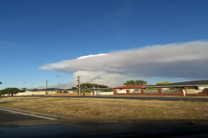Ein Auto fährt die Straße entlang und stößt eine große Rauchwolke aus, umgeben von Gras, Häusern, Bäumen, Masten und Laternen unter einem bewölktem Himmel.