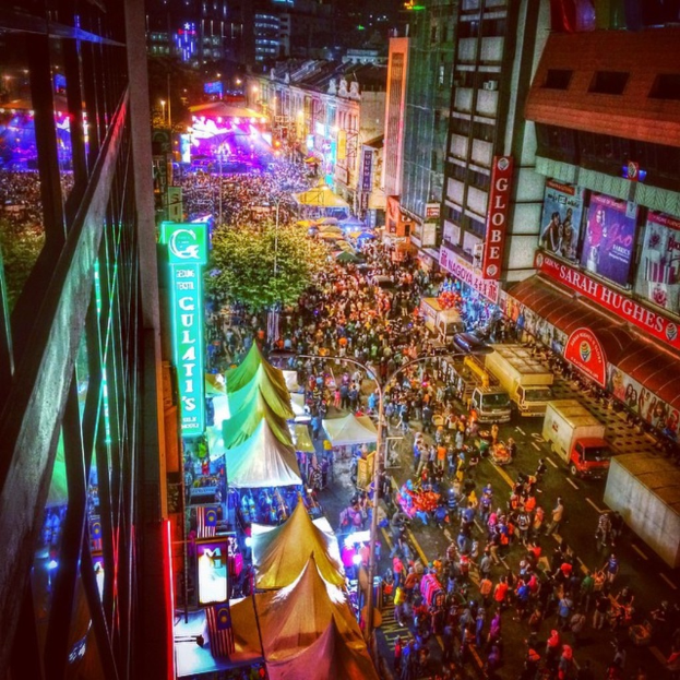 Eine volle KarnevalsSzene mit Zelten, Geschäften, Gebäuden und Bannern, mit hell beleuchteter Fläche im Hintergrund.