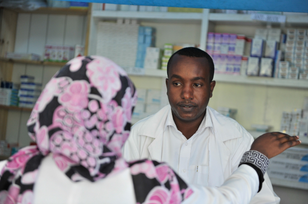 Eine Apothekerin in einem weißen Kittel steht einer sitzenden Frau in einer Apotheke gegenüber, mit Regalen voller Schachteln und Gegenständen im Hintergrund.