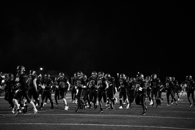 Schwarzes Bild von Fußballspielern in Helmen, die nachts auf das Feld laufen, mit Stadionlichtern und Pfosten im Hintergrund.