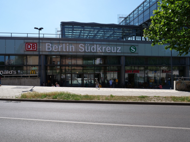 Großes glaswandiges Berliner Südkreuz-Bahnhof mit Text an seiner Fassade, umgeben von Straßenlaternen, Lampen, Fahrzeugen, Fußgängern, Fahrrädern, Bäumen und einem klaren blauen Himmel.