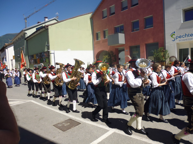 Eine Gruppe von Menschen in traditioneller bayrischer Tracht, die auf der Straße Musikinstrumente spielen und dabei durch eine Straße mit Gebäuden gehen, einige halten Fahnen, im Hintergrund ein Hügel und ein blauer Himmel.