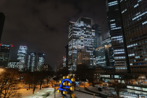 Das Hauptquartier der Europäischen Zentralbank in Frankfurt, Deutschland bei Nacht, mit beleuchteten Gebäuden, Bäumen, Straßenlaternen, Straßenlaternen und Menschen, die auf der Straße unter einem bewölkten Himmel gehen, mit einem Wasserzeichen auf dem Bild.