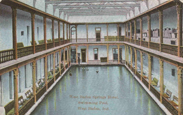 Eine alte Postkarte zeigt ein Hallenbad im West Baden Springs Hotel mit schwimmenden Menschen, umgeben von Säulen, Geländern, Stühlen und architektonischen Elementen sowie Text am unteren Rand.