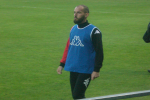 Ein Mann in einem blauen und roten T-Shirt steht auf einem Fußballfeld mit ausgestreckten Armen und entschlossener Miene, mit einem Metallstab unten und Gras im Hintergrund.