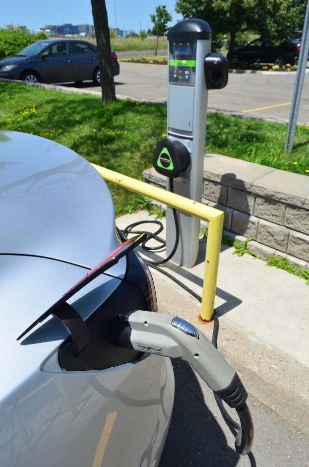 Ladestation für Elektroauto am Straßenrand mit Gras, Bäumen, Strommasten, Gebäuden und einem klaren Himmel im Hintergrund.