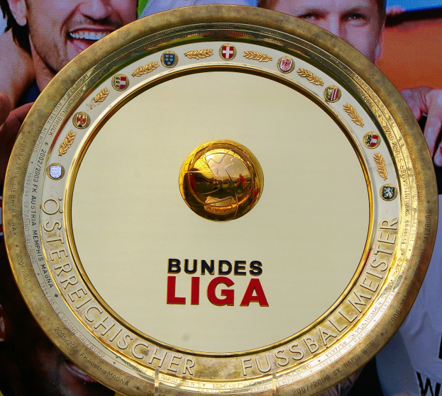 Ein Mann hält ein goldenes Schild mit der Aufschrift "Bundesliga" hoch, mit einem Banner im Hintergrund, auf dem drei Personen zu sehen sind.