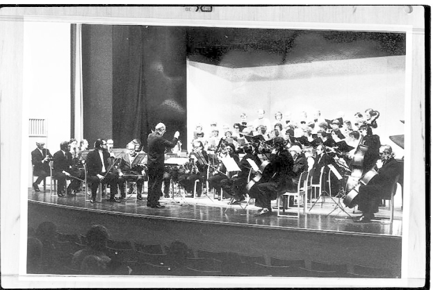 Ein Schwarz-Weiß-Foto eines Dirigenten in Anzug und Krawatte, der auf einer Bühne steht und ein Orchester mit Geigen, Celli und Flöten leitet, während das Publikum auf Stühlen sitzt und aufmerksam zuhört.