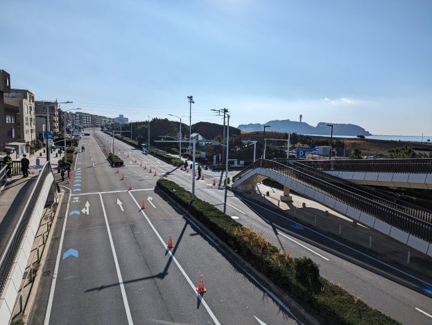 Straße mit Verkehrskegeln, eine Brücke mit Geländern, Laternenpfähle, Gebäude mit Fenstern und einige Menschen, vor Bergen und einem bewölkten Himmel, Teil des Takayama-Brückenprojekts.