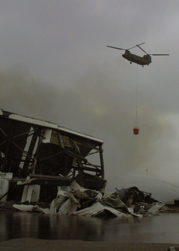 Ein Hubschrauber mit einem Seil fliegt in der Luft und lässt Wasser auf ein teilweise eingestürztes Gebäude regnen, mit dem Himmel im Hintergrund.