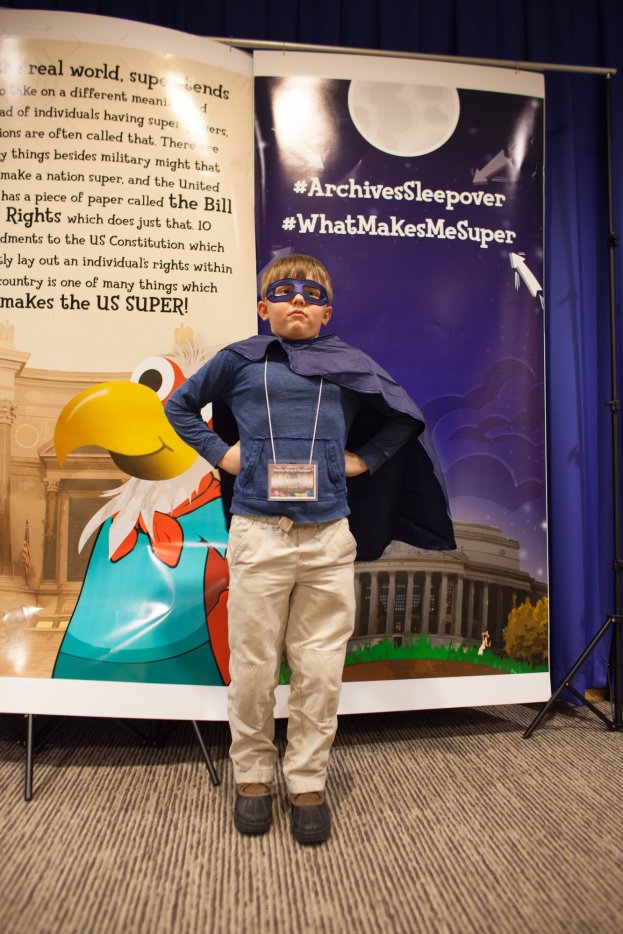 Junge in Superheldenkostüm vor einem Banner mit Text und einem Vogelbild, trägt Schuhe und hat einen Vorhang im Hintergrund.