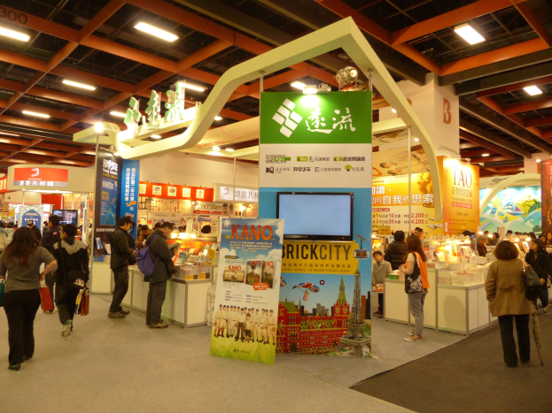 Eine Gruppe von Menschen vor einem Messestand mit Bannern, Fernseher, Tischen mit Gegenständen, Schildern und Deckenleuchten.