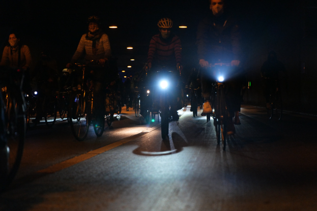 Eine Gruppe von Menschen, die nachts mit Fahrrädern auf einer gut beleuchteten Stadtstraße fahren, alle tragen Helme.