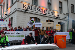 Eine Gruppe von Menschen steht vor einem Gebäude und hält Schilder und Plakate hoch, während zwei Personen im Vordergrund sitzen und ein Müllcontainer rechts zu sehen ist, bei einer Demonstration in Deutschland, mit Gebäuden im Hintergrund, die Fenster, Lichter und Schilder aufweisen.