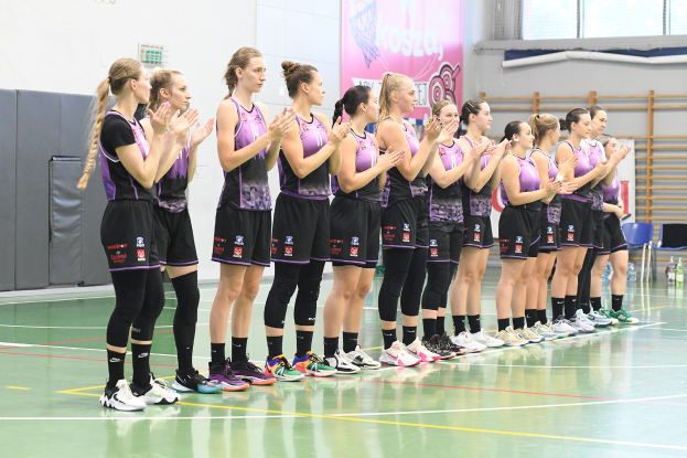 Eine Gruppe von Frauen steht auf einem Basketballfeld, klatscht begeistert, mit einem Banner im Hintergrund, das 'Koszia - Frauen-Handball-Team' lautet