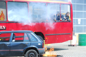 Roter Doppeldeckerbus mit Rauch und drei sichtbaren Passagieren, neben einem Auto geparkt, mit einem Glasfenster-Gebäude und einem Fass im Hintergrund.