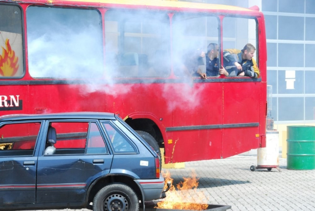 Roter Doppeldeckerbus mit Rauch und drei sichtbaren Passagieren, neben einem Auto geparkt, mit einem Glasfenster-Gebäude und einem Fass im Hintergrund.