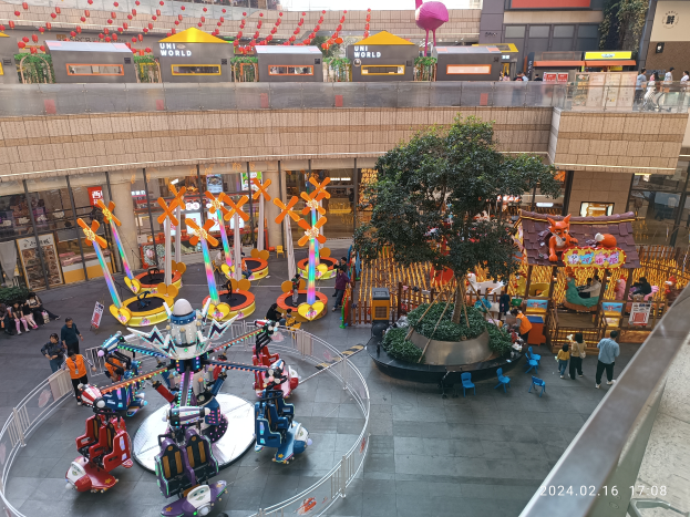 Ein Vergnügungspark innerhalb eines Einkaufszentrums mit verschiedenen Fahrgeschäften, Bäumen, Menschen und Ständen, mit Gebäuden oben und einem Wasserzeichen unten rechts.