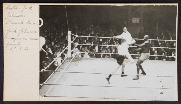 Schwarzes und weißes Foto von zwei Boxern, Jack Johnson und Frank Morgan, in einem Boxring mit Zuschauern im Hintergrund.