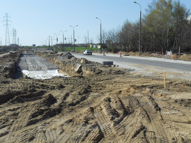 Eine Baustelle mit einem großen Haufen Erde neben einer Straße, Fahrzeuge fahren, Laternenpfähle, Strommasten mit Drähten, Bäume und einen klaren blauen Himmel.