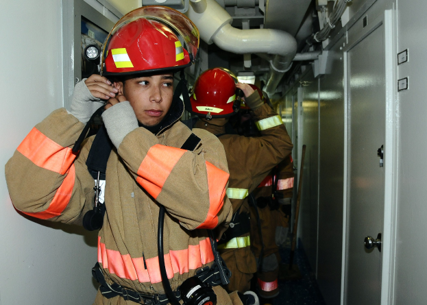 Gruppe von Feuerwehrmännern in Uniform, die zusammen in einem Trainingsraum mit einer Tür auf der rechten Seite und einer Wand auf der linken Seite stehen.