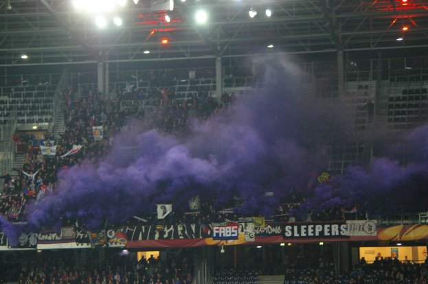 Große Menschenmenge in einem Stadion mit violettem Rauch, Bannern, Stühlen, Treppen, Geländern und Deckenlampen.