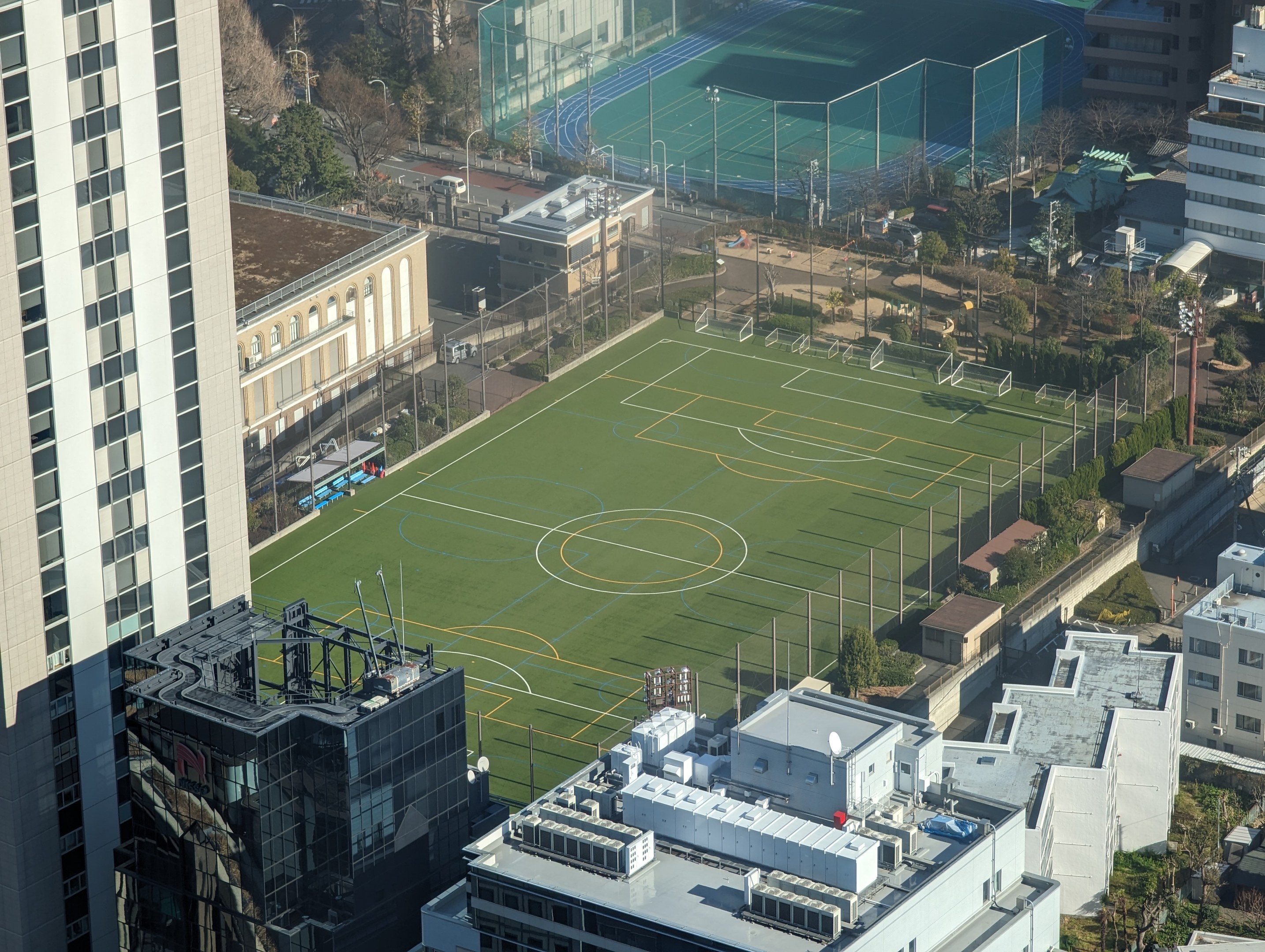 Luftaufnahme eines Fußballfelds in Tokyo, Japan, umgeben von Stadtgebäuden, Bäumen, Fahrzeugen und einem Zaun.