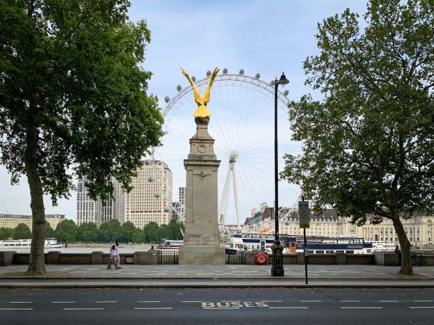 Das London Eye Riesenrad in London mit einer Statue auf einem Podest im Vordergrund, eine Straße, Bäume, ein Lichtmast und Menschen, die auf einem Gehweg spazieren gehen, mit Schiffen, Gebäuden und Bäumen im Hintergrund.