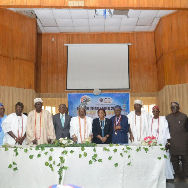Gruppe von Menschen um einen weißgedeckten Tisch versammelt, der mit Blumen und Blättern geschmückt ist und im Hintergrund ein Banner mit der Aufschrift "ICCI International Trade Fair 2019" sowie ein Projektionsscreen und Deckenbeleuchtung.
