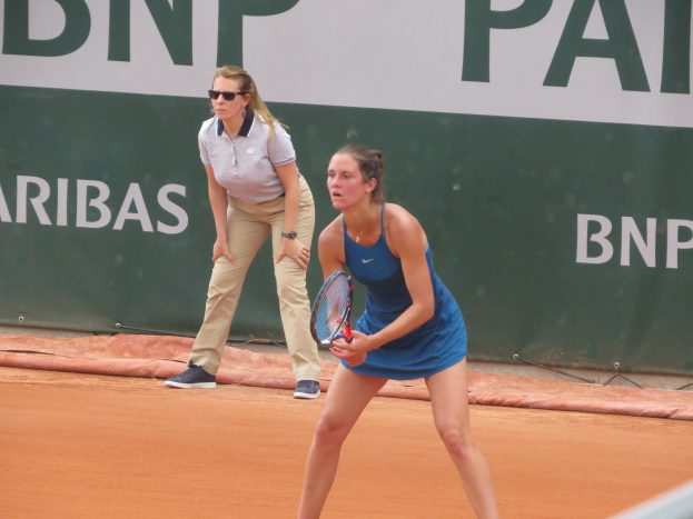 Zwei Frauen beim Tennis auf einem Sandplatz, wobei eine in einem blauen Kleid mit einem Schläger in der Hand im Vordergrund steht und eine andere dahinter steht, während ein grüner Banner mit Text im Hintergrund erscheint.