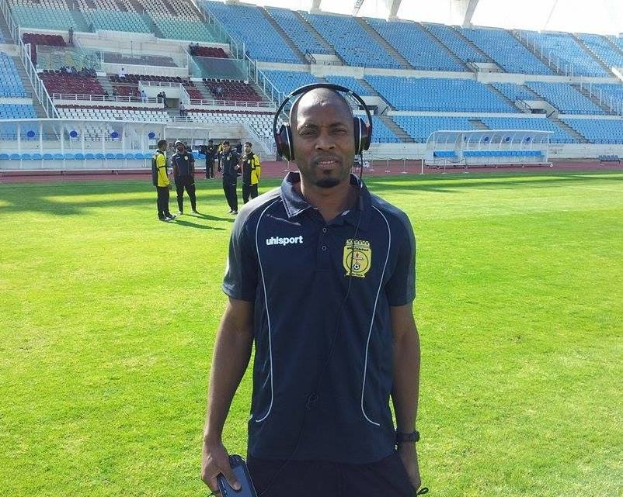 Ein Mann in einem Headset, wahrscheinlich der Trainer der ghanaischen Nationalmannschaft, steht auf einem Fußballfeld neben einem Stadion, hält ein Objekt und hat ein paar Menschen im Hintergrund und den Himmel über sich.