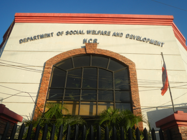 Das Gebäude des Departments für Soziales und Entwicklung mit Text an der Wand, einem Fahnenmast mit einer Flagge, einem Metallzaun, Pflanzen, Bäumen, Drähten und einem bewölkten Himmel.