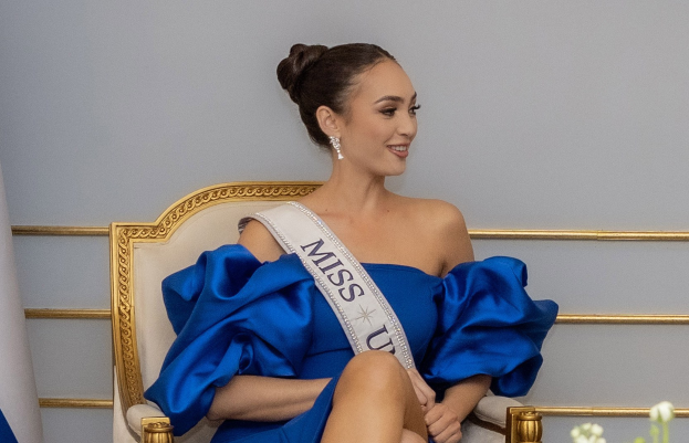 Frau in einem blauen Kleid mit einer "Miss Universe 2019"-Schärpe, die auf einem Stuhl sitzt und von Blumen umgeben ist.