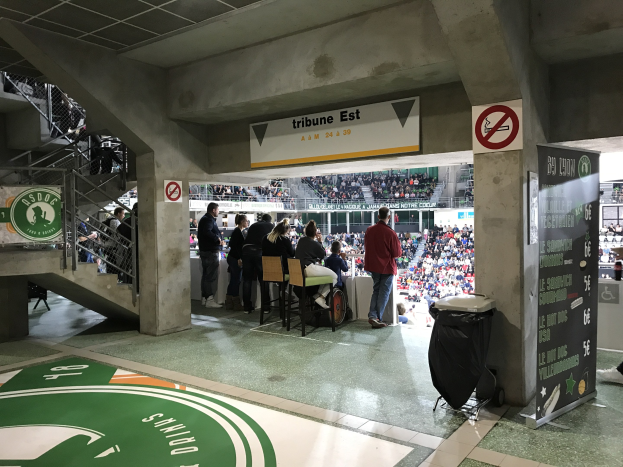 Eine Gruppe von Menschen steht vor einer sitzenden Menge in einem Stadion während eines Boston Celtics vs New York Jets-Spiels, mit Schildern, einem Müllcontainer, einer Treppe und Deckenleuchten.