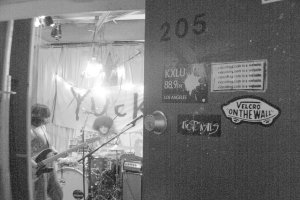 Ein Schwarz-Weiß-Bild eines Mannes, der vor Trommeln sitzt, einer Frau, die Gitarre spielt, und einem Banner mit der Aufschrift 'YUCK' an einer Tür.