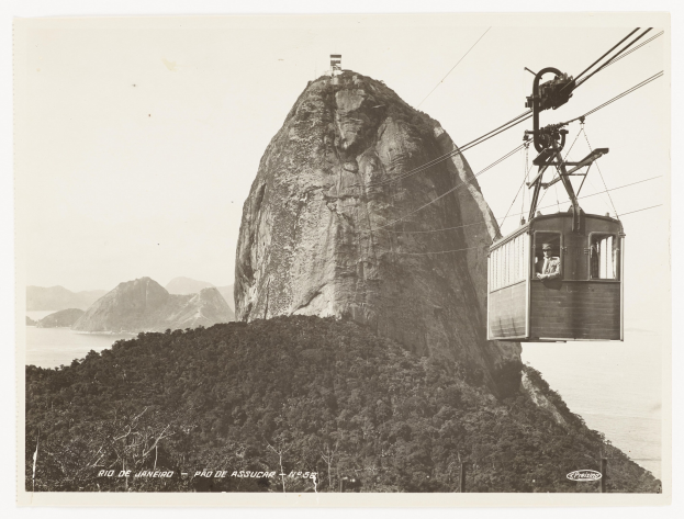 Schwarz-weiß-Foto einer Seilbahn, die einen Berg hinauffährt, mit einer Person darin, umgeben von Bäumen, Hügeln und Wasser; Text unten.