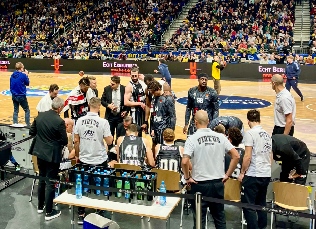 A group of men stand on a basketball court surrounded by spectators, with a person holding a camera on the left and boards with text in the background.