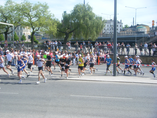Läufer bei einem Marathon überqueren eine Ziellinie, die mit einem Band an einem Pfahl markiert ist, auf einer Straße, die von einem Metallzaun, einem Torpfosten, einem an einem Pfahl befestigten Band, einigen Menschen auf dem Gehweg, einer Metallabsperrung, einigen Pfosten, Straßenpfosten, Schildern, einer Brücke, einigen Gebäuden mit Fenstern, Bäumen und einer bewölkten Himmel umgeben ist.