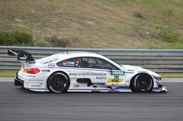 BMW M4 DTM-Rennwagen auf einer Rennstrecke mit grünem Rasen und einer Absperrung im Hintergrund.