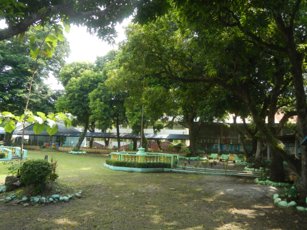 Ein Park mit saftigem Grün, Pflanzen und Bäumen, der von einem zentralen Brunnen dominiert wird, mit Gebäuden und einem klaren blauen Himmel im Hintergrund.