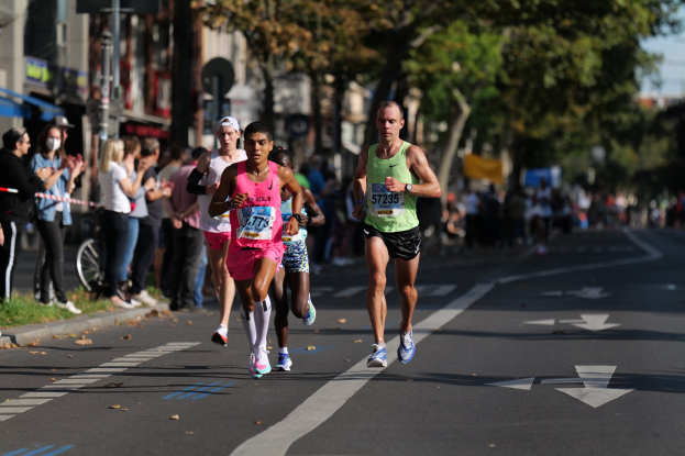 Gruppe von Läufern bei einem Stadtmarathon, mit Zuschauern und städtischen Elementen wie Bäumen, Gebäuden und einem Fahrrad im unscharfen Hintergrund.