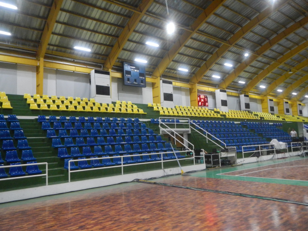 Großer Hallensportplatz mit Holzboden, blauen und gelben Sitzplätzen, Geländern, Treppen, wandmontierten Bildschirmen, Deckenleuchten und einigen anwesenden Personen.