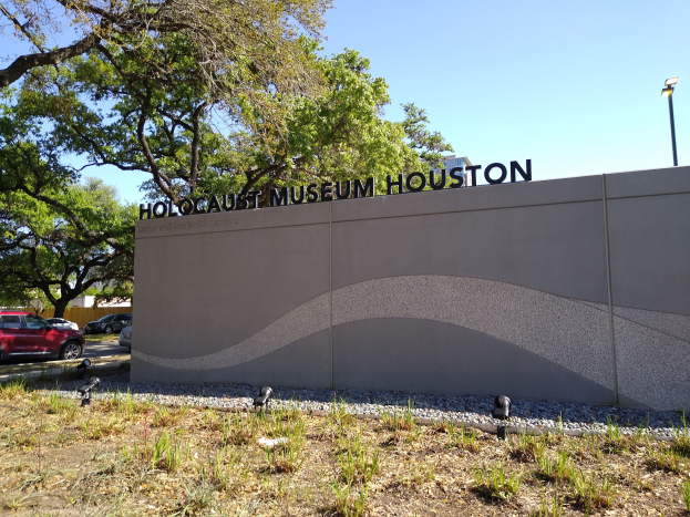 Außensicht des Holocaust-Museums Houston mit eingravierter Textwand, Gedenksteine, Landschaft, Straße mit Fahrzeugen, Straßenmast und bewölktem Himmel.