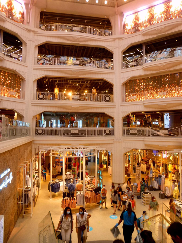 Ein belebtes Einkaufszentrum mit Menschen, die durch die Gänge schlendern, Taschen tragen und Schaufensterpuppen mit Kleidern ansehen, umgeben von Geländern, Beleuchtung und Wandtexttafeln.