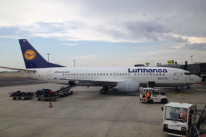 Ein Lufthansa Airbus A320-200 steht auf dem Frankfurter Flughafen, umgeben von verschiedenen Bodenfahrzeugen, Wagen, Absperrungen, Pfählen und Texttafeln unter einem bewölkten Himmel.