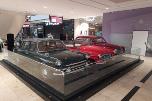 Eine Autohalle mit einer Glasvitrine voller Oldtimer, umgeben von Blumenbouquets, Namensschildern, Deckenleuchten und anderen Gegenständen.