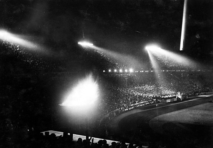 Schwarzes und weißes Foto eines vollen Stadions, das von Flutlicht gegen einen dunklen Himmel beleuchtet wird.