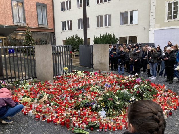Eine Gruppe von Menschen versammelt sich um ein provisorisches Denkmal mit Blumen und Kerzen vor einem Gebäude, während eine Person links kniet und eine Kamera hölt, mit Gebäuden und einem Tor im Hintergrund.