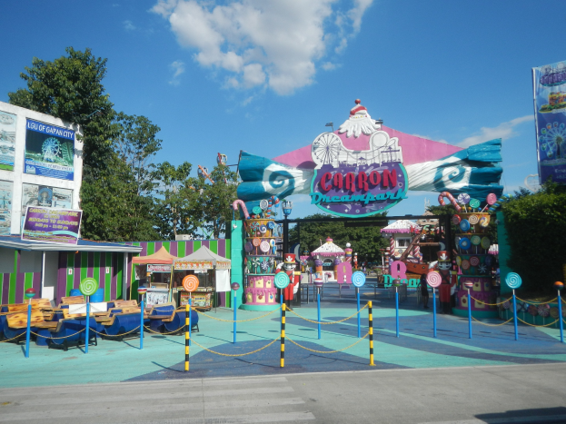 Karussell am Eingang eines Parks mit einer Straße, einem Zaun, Buden, Schildern, Bäumen und einem bewölkten Himmel im Hintergrund.